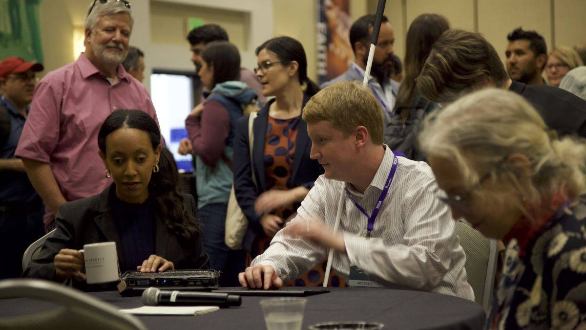 A man with a white cane and two women seated at a table in a crowded event hall at AccessU.