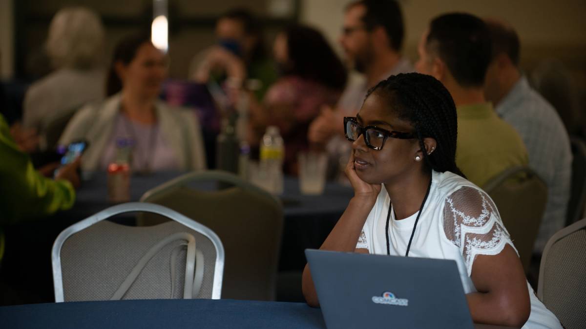 woman seated at a table with a laptop listening to a speaker