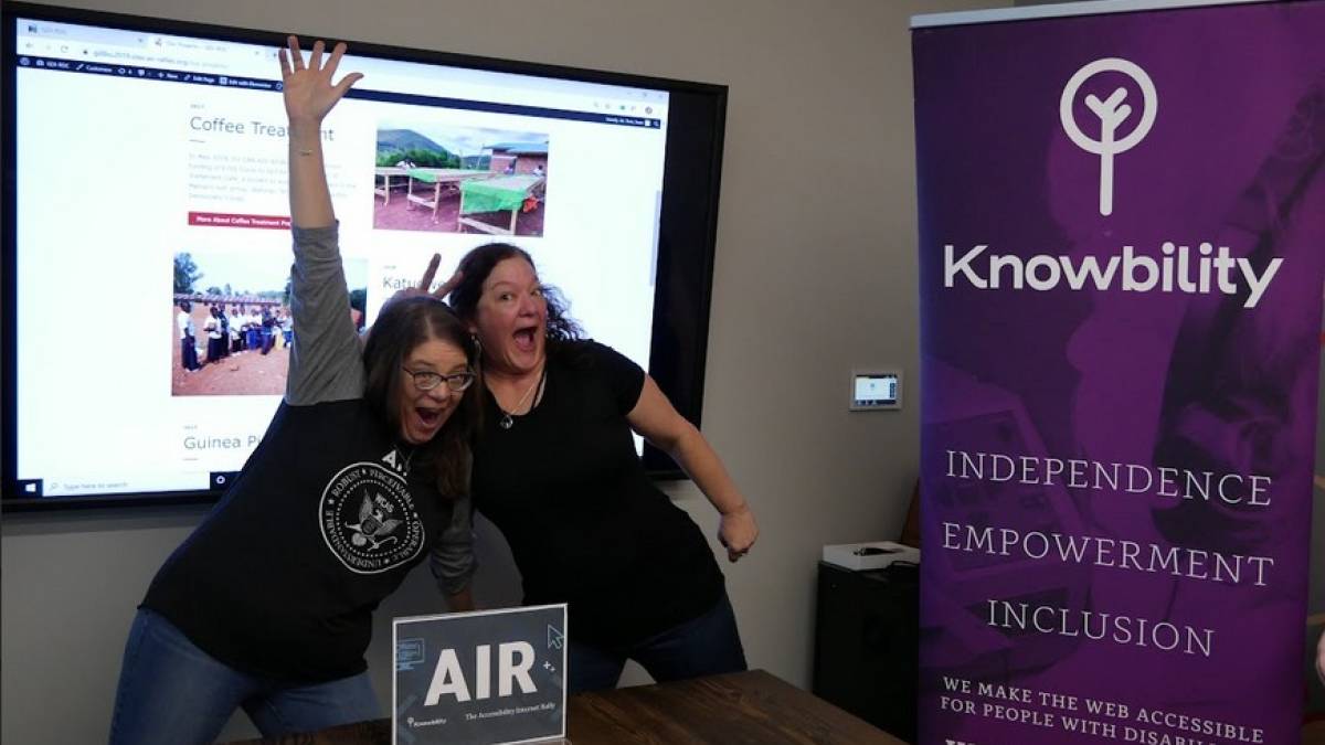 Two excited AIR participants pose before a large screen of one the AIR sites, and AIR sign and a Knowbility banner.
