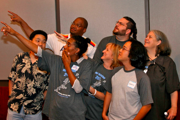 AIR Austin 2007 team photograph featuring seven team members, including Sharron Rush, Knowbility's executive director. The group is captured in a lively moment, with several individuals pointing to the left, one person covering their mouth in surprise, and two members sharing chuckles. The dynamic scene suggests a sense of curiosity and shared amusement, portraying the team's engaging interaction and camaraderie.