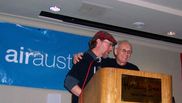Jim Allan and Jim Thatcher on stage behind a podium and microphone. Jim Thatcher, on the right, is smiling and has his arm around Jim Allan’s shoulder in a friendly and supportive pose, capturing a warm moment at AIR Austin 2008.