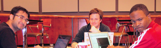 Three people seated around a table, laptops in front of them. The central and left individuals wear smiles, while on the right, a playful expression is evident, with an arched eyebrow adding a touch of light-heartedness. Their collaborative atmosphere reflects a productive and enjoyable teamwork.