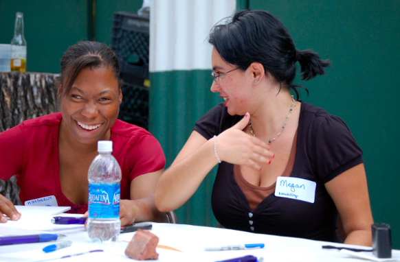 Two people seated at a table, engaging in conversation. The person on the right speaks animatedly, while the person on the left wears a bright smile, exuding happiness and laughter.