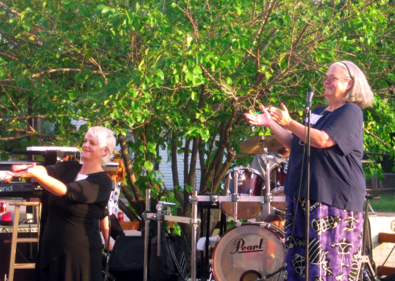 Kicking off AIR Austin Rocks in 2010, two figures stand on a stage against a backdrop of musical instruments. Engaged in enthusiastic applause, Sharron Rush, Knowbility's executive director, stands on the right. On the left, an ASL interpreter enhances accessibility. The scene captures a moment of shared joy and inclusivity, setting the tone for the event's spirited beginning