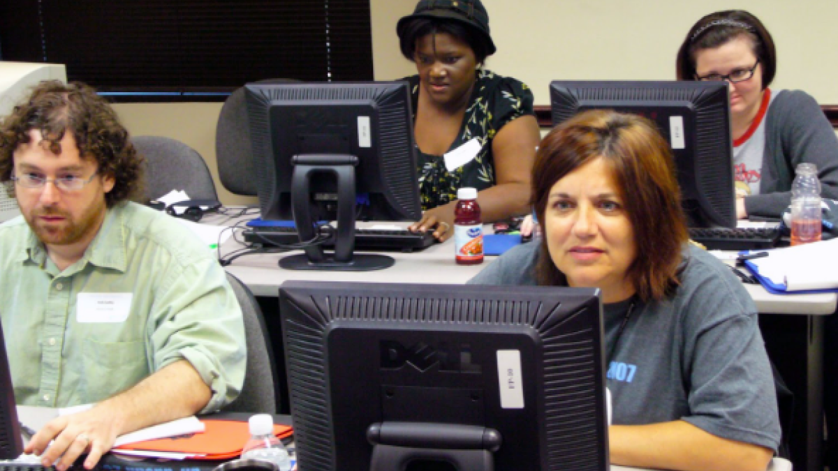 Four people sitting at a table, each focused on their own computer, engaged in work. The atmosphere is collaborative, with a sense of concentration and productivity.