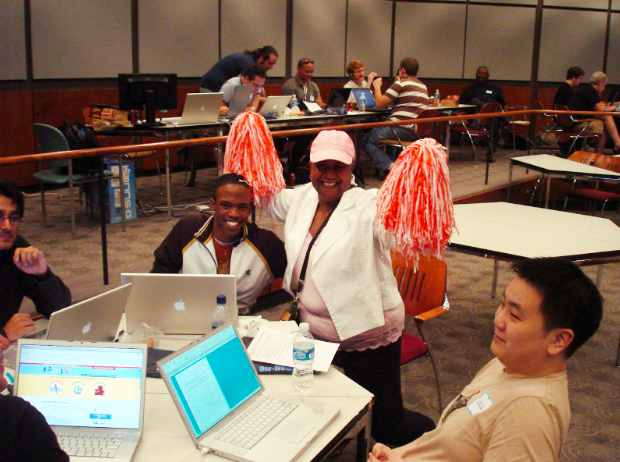 Scene from AIR- A conference room filled with participants. At the heart of the image, two individuals pose for the camera while at a table surrounded by colleagues with laptops. One person leans over with a smile towards a standing person beside them, who enthusiastically waves cheerleader pom-poms in the air, creating an atmosphere of energetic camaraderie.