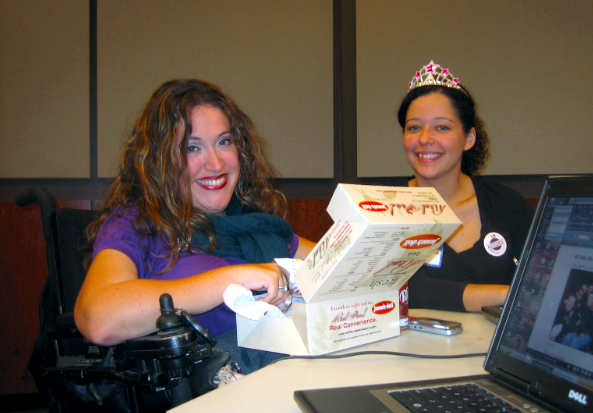 Two individuals seated at a table with laptops sharing joyful smiles. On the left, a person retrieves a sandwich from a box, while on the right, another person wears a plastic tiara, adding a touch of whimsy and lightheartedness to the scene.