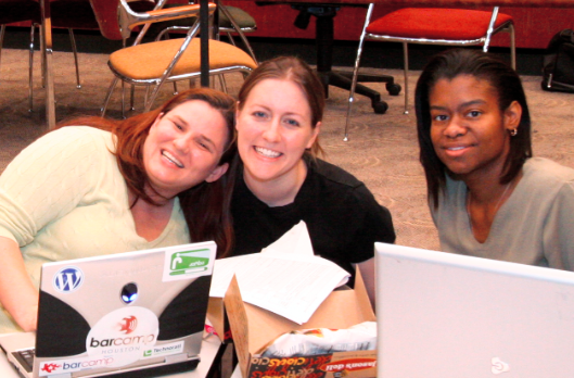 Three individuals at a table, sitting in front of laptops, wearing smiles that radiate a sense of camaraderie and collaboration.