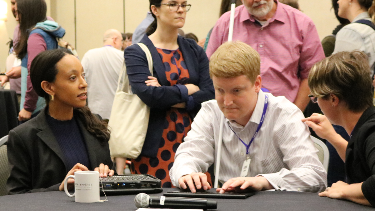Conference attendees talking at a table together. One is using a braillenote and another is using a tablet and has a white cane propped on thier shoulder