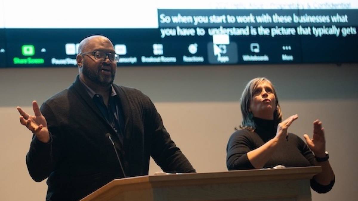 AccessU 2022 keynote speaker Kelsey Ruger behing the podium with an ASL interpreter beside him.