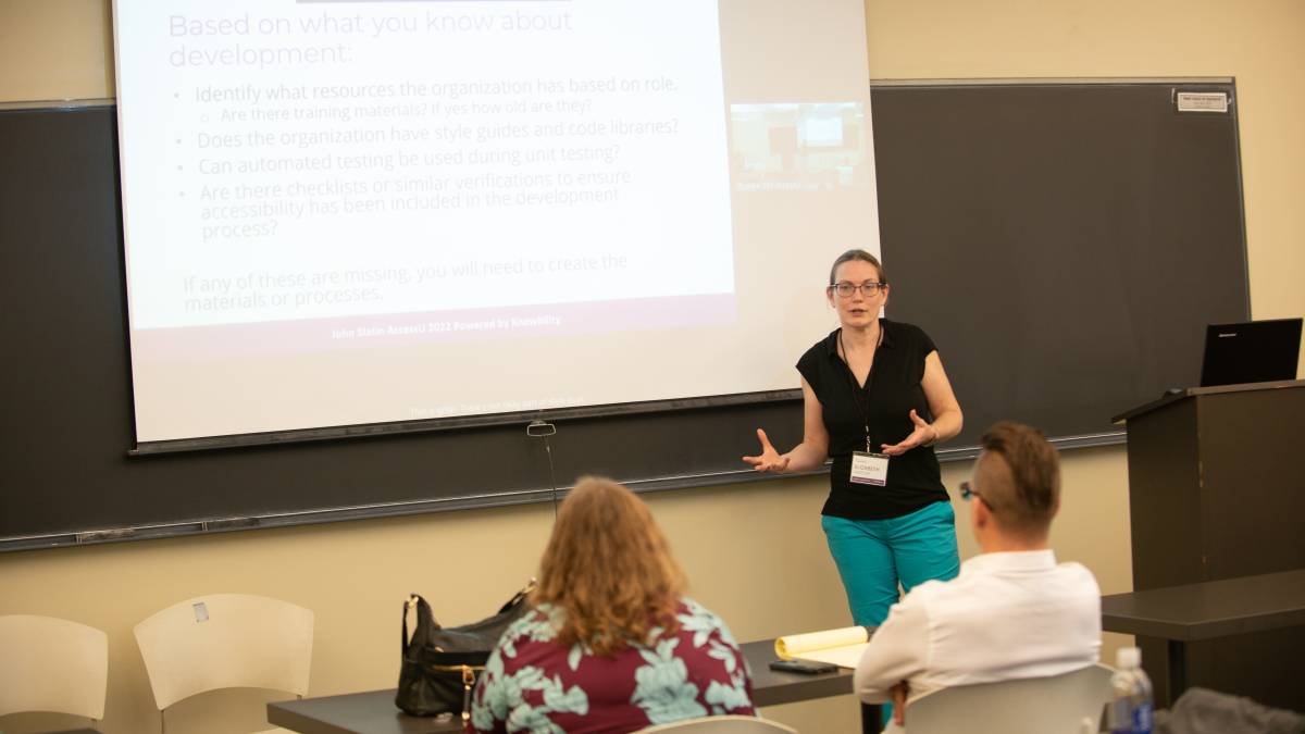 AccessU 2022 presenter in a classroom with attendees, talking in front of screen displaying session content and zoom participants