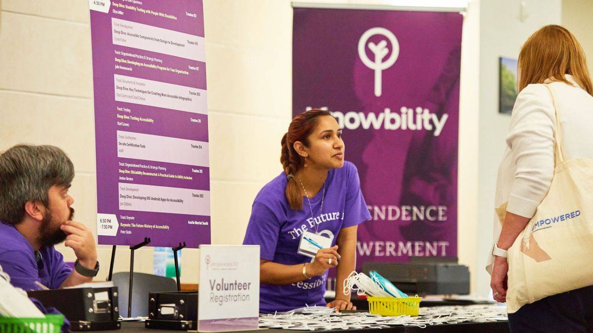 One volunteer at AccessU 2024 registration desk assists an attendee while another volunteer looks on.