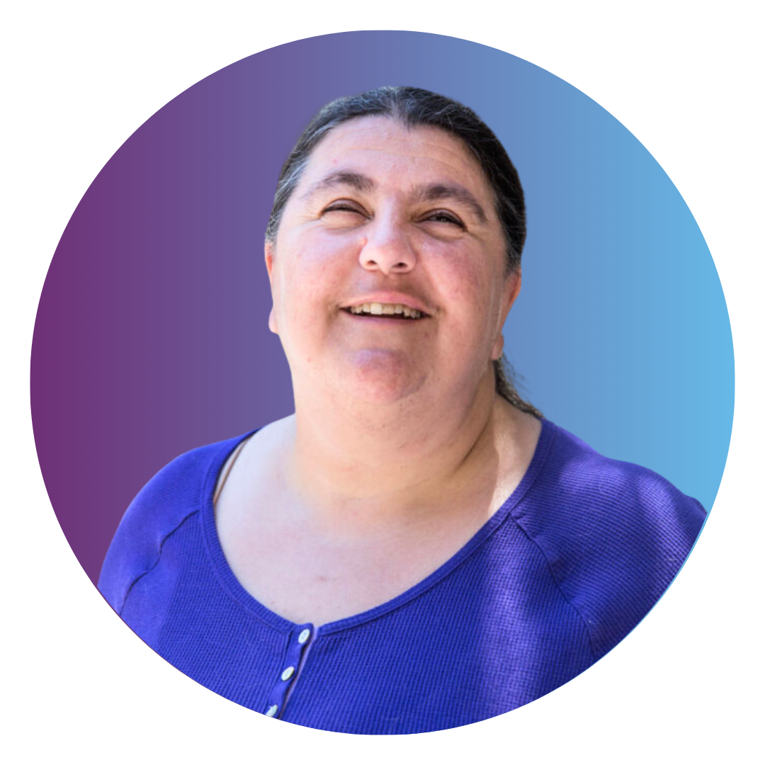 A smiling woman with long salt-and-pepper black hair tied back in a ponytail. She wears a blue shirt with white buttons.