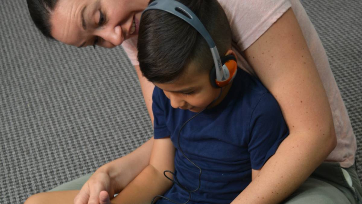 A boy using headphones and a tablet, seated in a woman's lap. She is looking at the tablet screen with him.