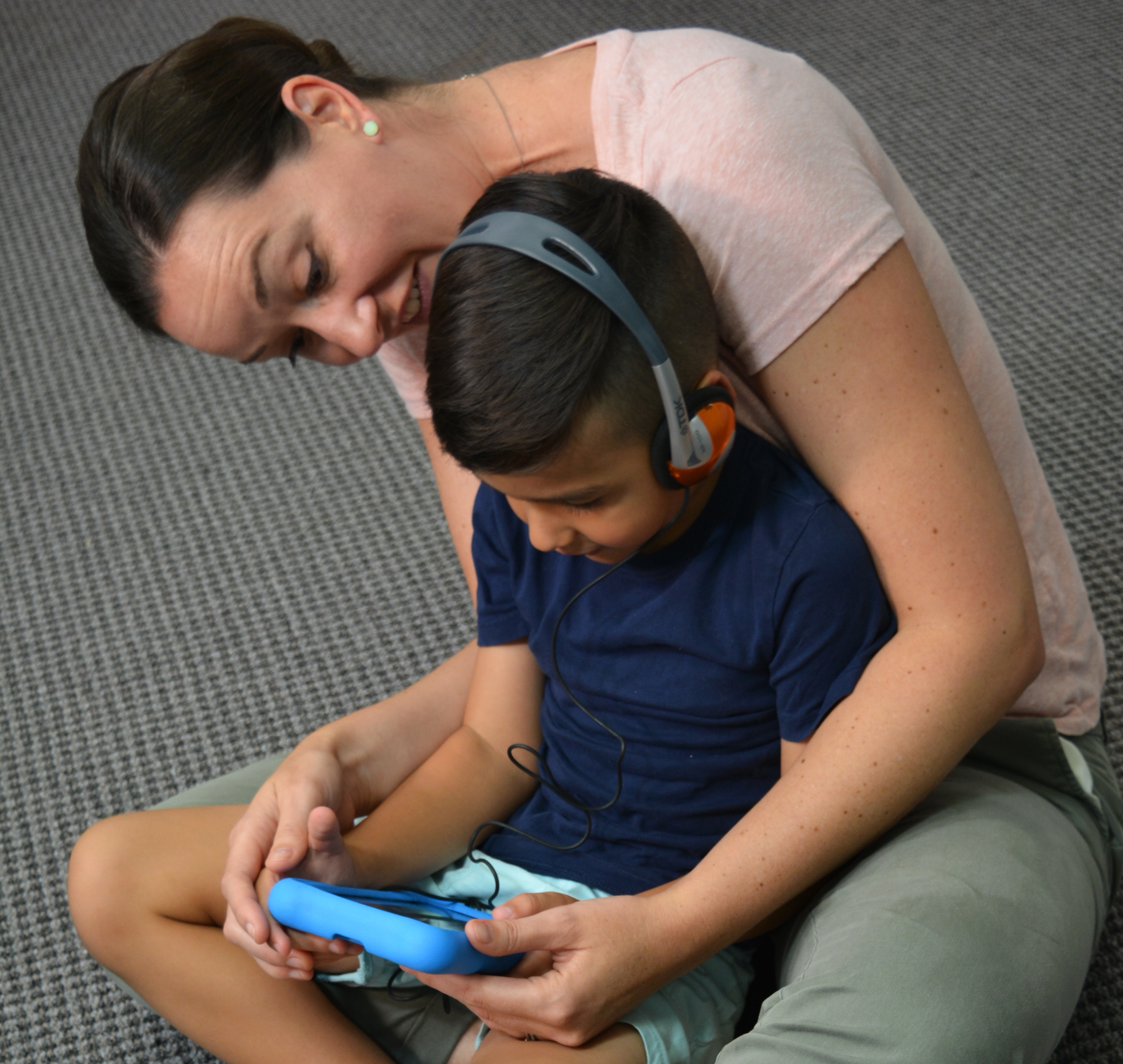 A boy using headphones and a tablet, seated in a woman's lap. She is looking at the tablet screen with him.